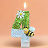 A golden glitter number four candle decorated with a daisy and a bee, lit by a flame.