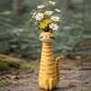 A yellow resin cat figurine with a flower vase on its head, set in a natural outdoor environment.