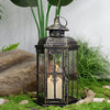 A vintage Nordic iron candle lantern with glass panes sits on mossy ground next to large rocks and greenery.