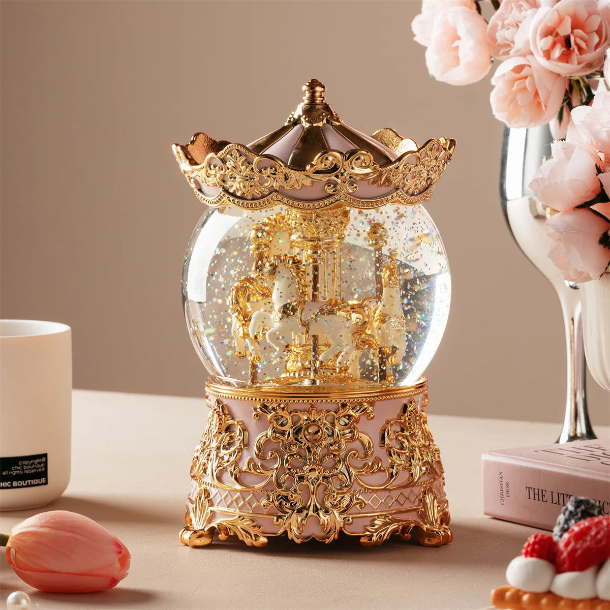 Adorno de caja musical de orbe de cristal decorativo con detalles dorados, junto a flores rosas y una fruta color melocotón