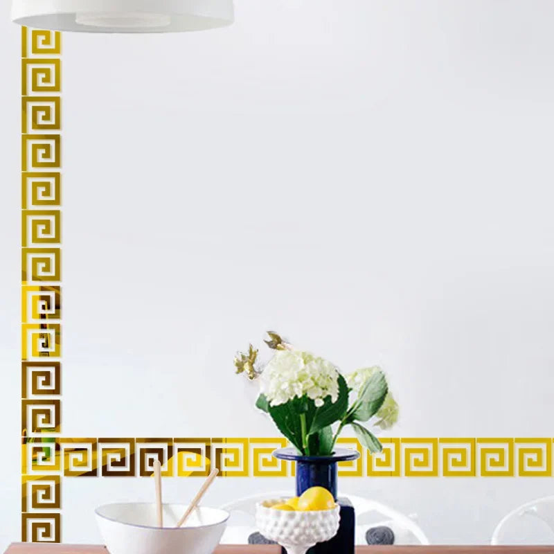 A decorative border of gold geometric tiles on a white wall, with a white bowl and green flowers in the foreground.