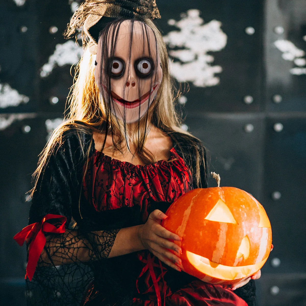 A woman wearing a full face horror costume with pale makeup and long hair holds an orange carved pumpkin.