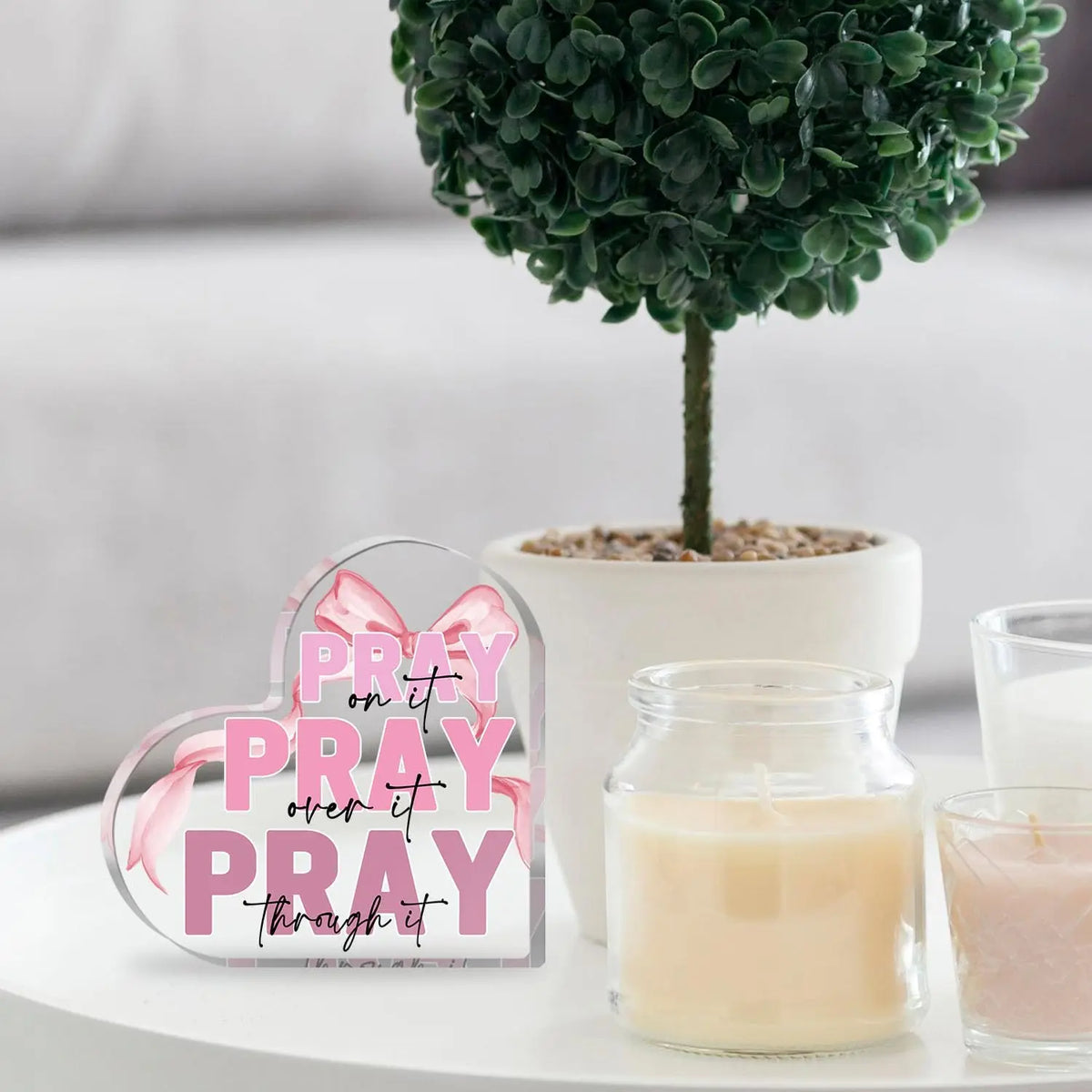 Pink acrylic heart sign with the text Pray on It Pray over it Pray through it displayed next to a candle and a green plant