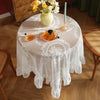 A round table set for a meal with an elegant white floral lace tablecloth, a vase of white flowers, and an orange glass.
