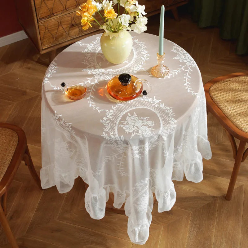 A round table set for a meal with an elegant white floral lace tablecloth, a vase of white flowers, and an orange glass.