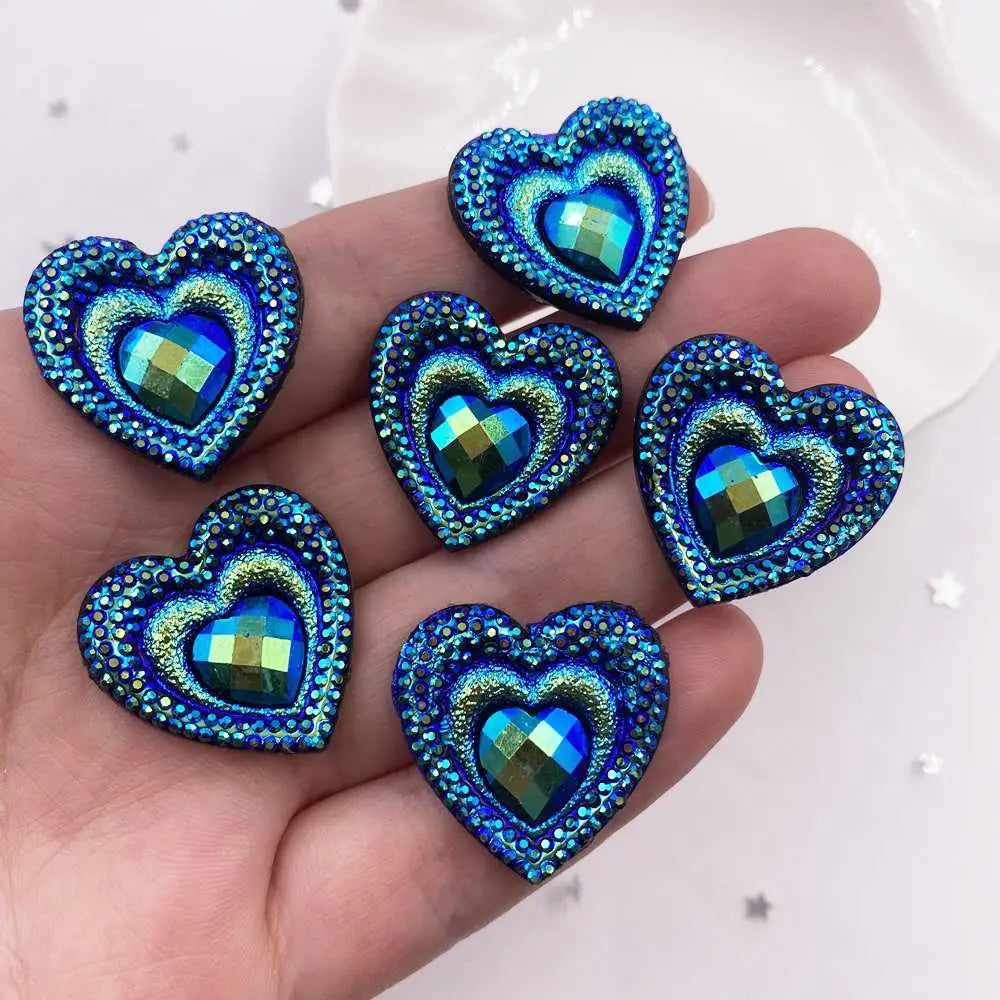 A hand holds a set of twelve heart-shaped blue and green iridescent peacock resin rhinestone flatbacks.