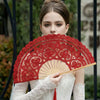 A woman holds a red lace hand fan with a tassel pendant against a blurred outdoor background.