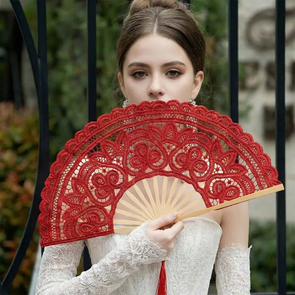 A woman holds a red lace hand fan with a tassel pendant against a blurred outdoor background.