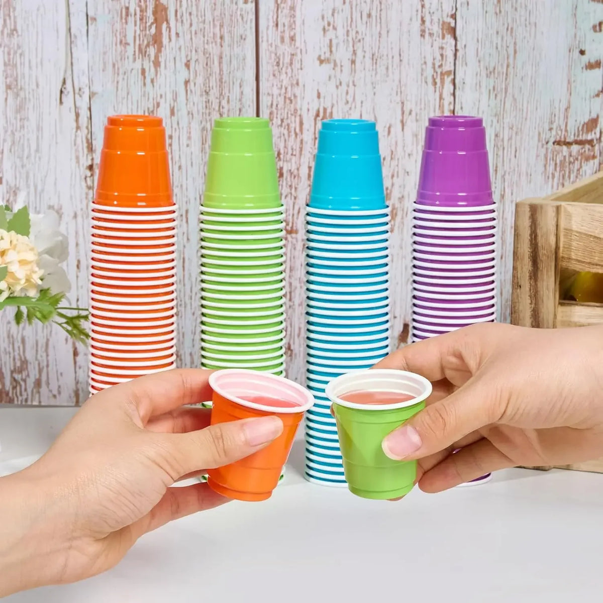 A hand holds a stack of colorful disposable plastic shot cups in various shades of orange, green, blue, and purple.
