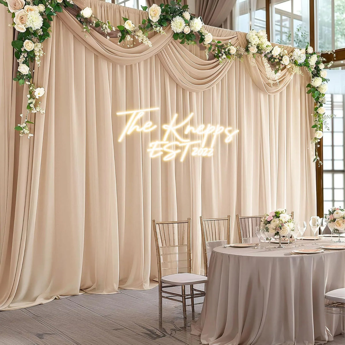 A beige chiffon backdrop curtain decorated with white floral garlands and a glowing script sign for an event.