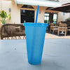 A bright blue glitter party cup with a straw is placed on a white table outdoors.