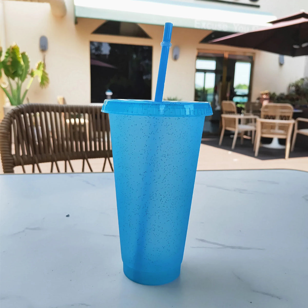 A bright blue glitter party cup with a straw is placed on a white table outdoors.