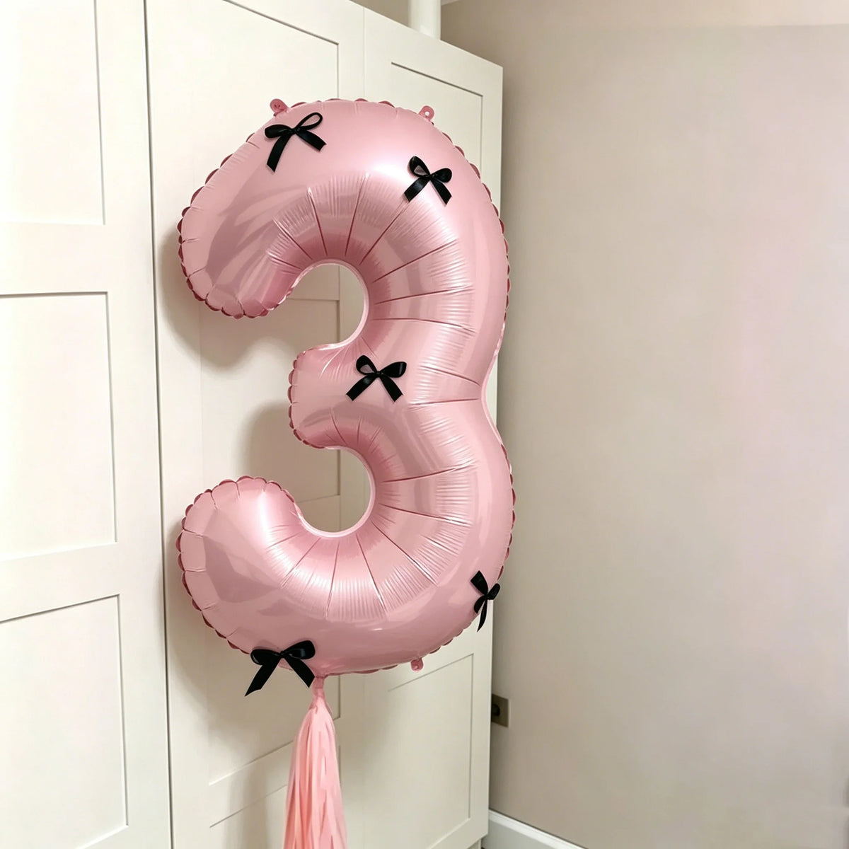 A pink foil balloon shaped like the number 3 with black bows, hanging on a white cabinet door.