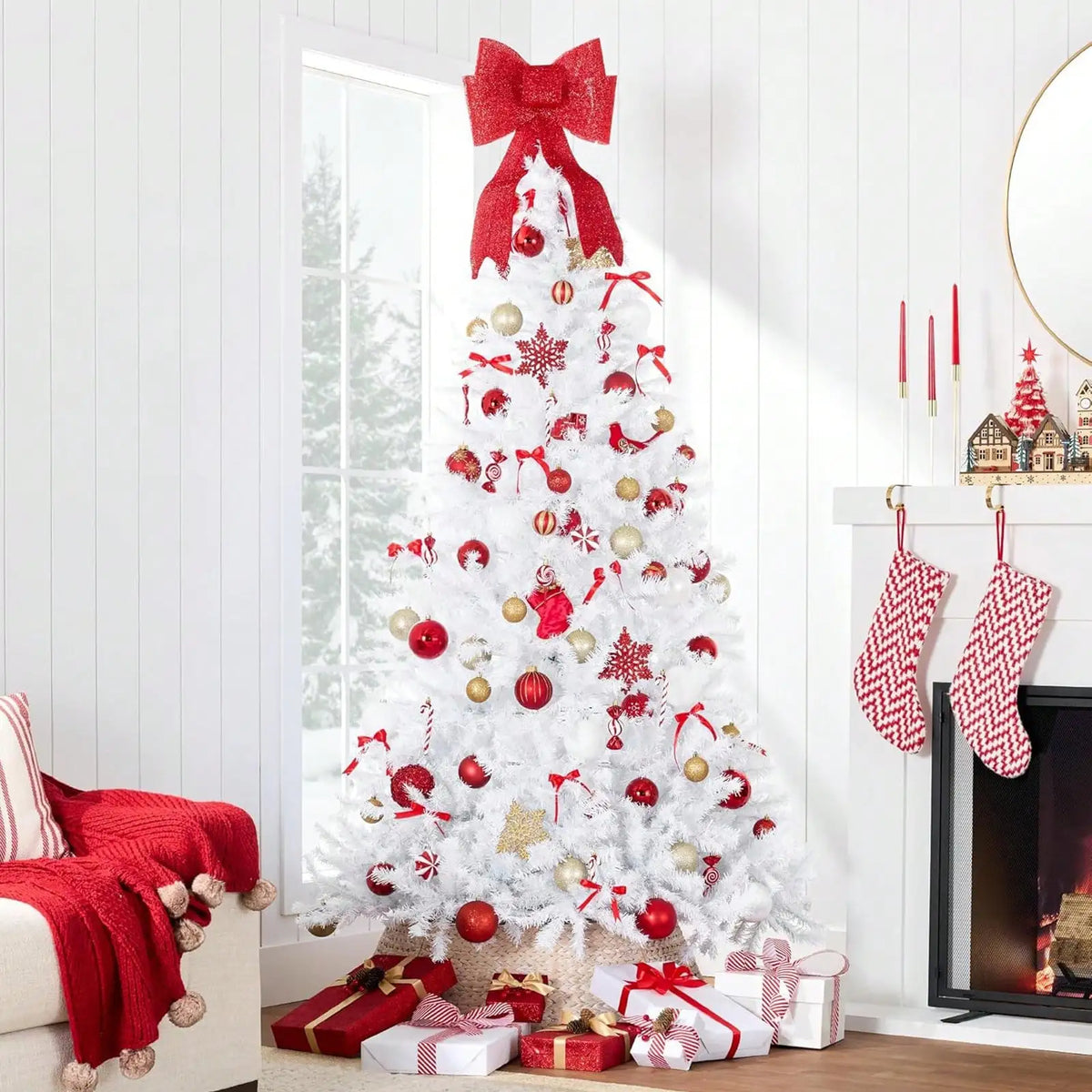 A white artificial Christmas tree decorated with red ornaments and bows, standing next to a fireplace.