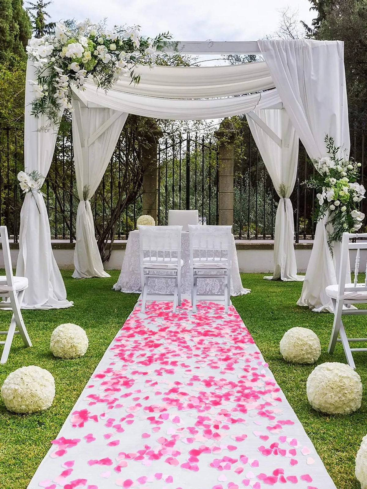 A wedding arch decorated with white draping and greenery, featuring a pink confetti aisle on a lawn.