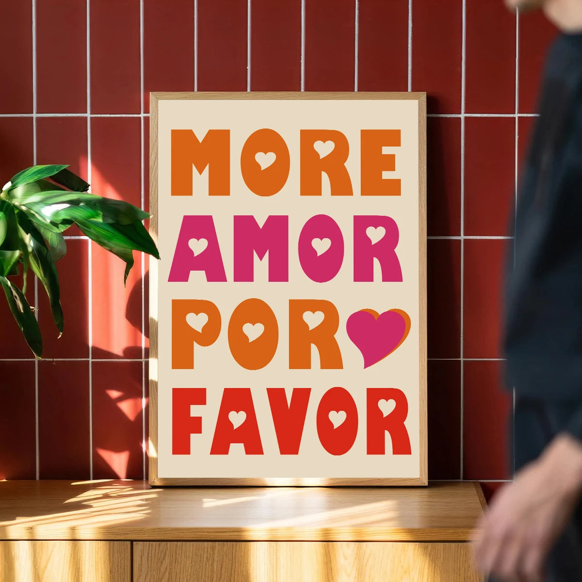 A framed canvas print featuring the text MORE AMOR POR FAVOR in orange and pink hearts on a light wood background.