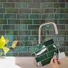 A hand is installing a square green peel and stick vinyl wall tile onto a dark teal subway tile backsplash.