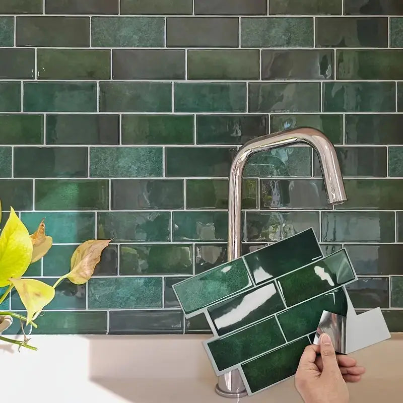A hand is installing a square green peel and stick vinyl wall tile onto a dark teal subway tile backsplash.