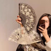 A woman holds an elegant lace rose folding fan with intricate floral patterns against a light background.