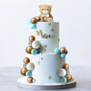 A two-tiered light blue cake decorated with gold and blue spherical candies and a bear topper.