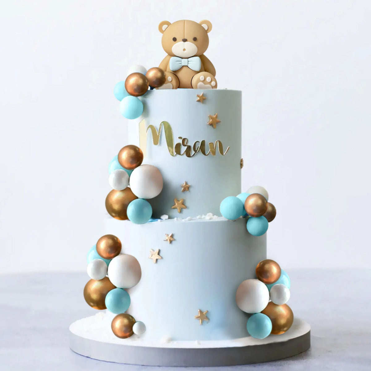 A two-tiered light blue cake decorated with gold and blue spherical candies and a bear topper.