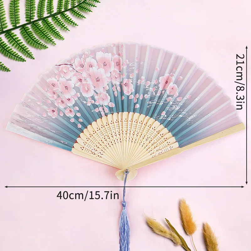 A delicate pink and blue floral bamboo folding hand fan with a tassel displayed on a light pink background.