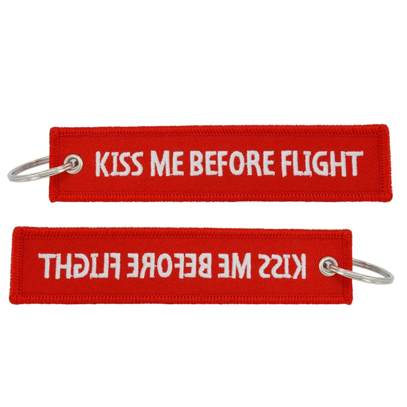 Red woven tag with white text that reads Kiss Me Before Flight and a metal keyring.