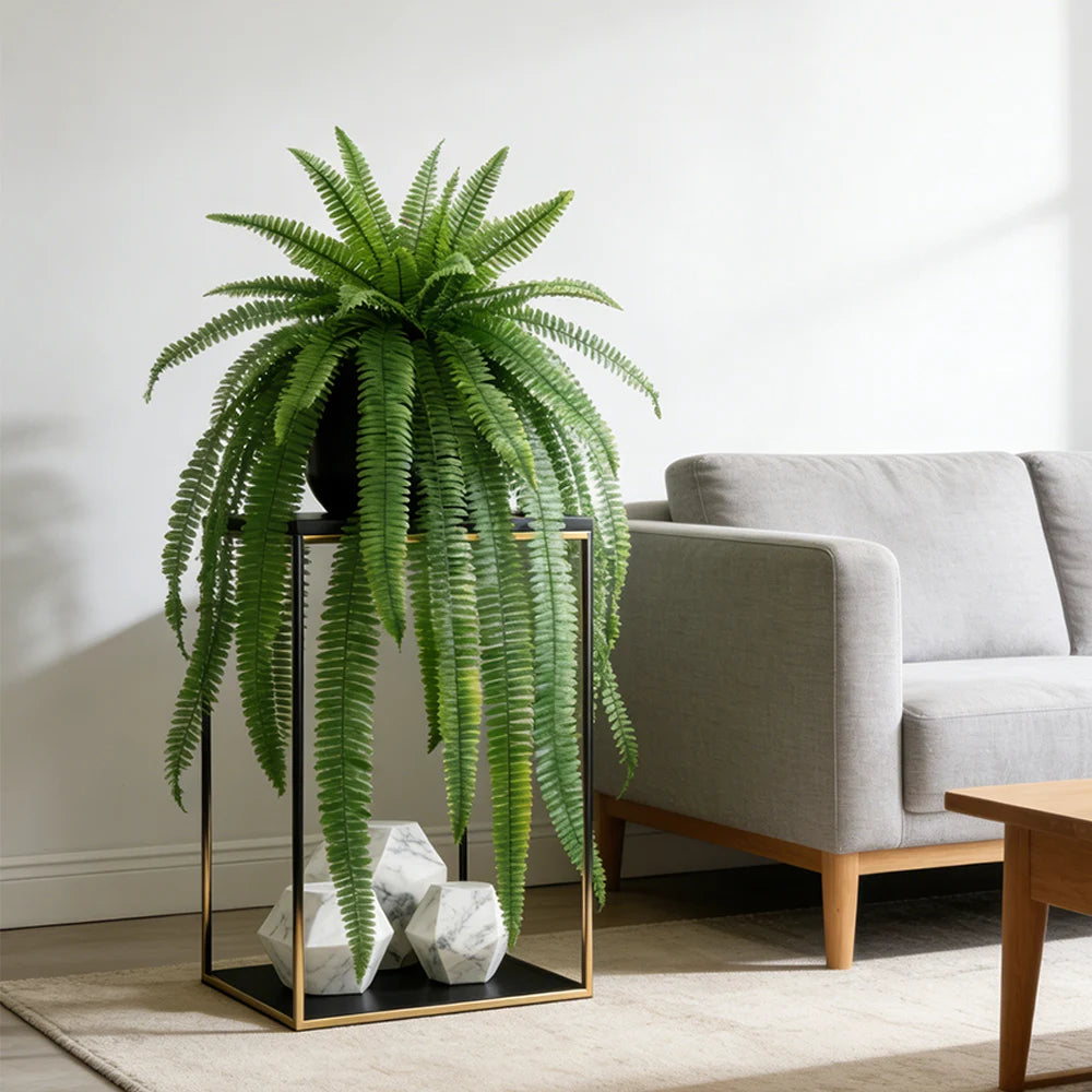 Artificial Persian fern hanging vine in a black pot displayed on a gold-framed stand next to a sofa.