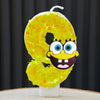 A yellow sponge cake candle shaped like the number 0, featuring a cartoon face.