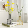 A black iron hollow vase holding yellow flowers next to a gold wire vase with green foliage.