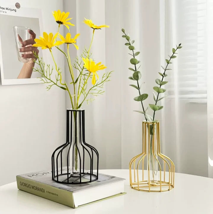 A black iron hollow vase holding yellow flowers next to a gold wire vase with green foliage.