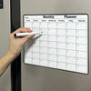 A hand writes on a monthly planner calendar displayed on a refrigerator door.