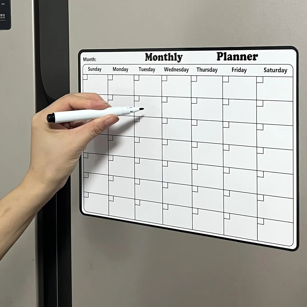 A hand writes on a monthly planner calendar displayed on a refrigerator door.