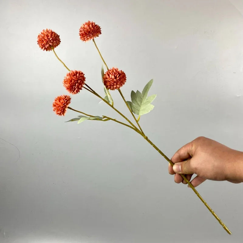 Una mano sostiene un tallo con varias flores artificiales de crisantemo y diente de león naranjas-rojas y esponjosas.