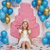 A young girl sits in front of a pink tiered cake photo backdrop decorated with gold and blue balloons.
