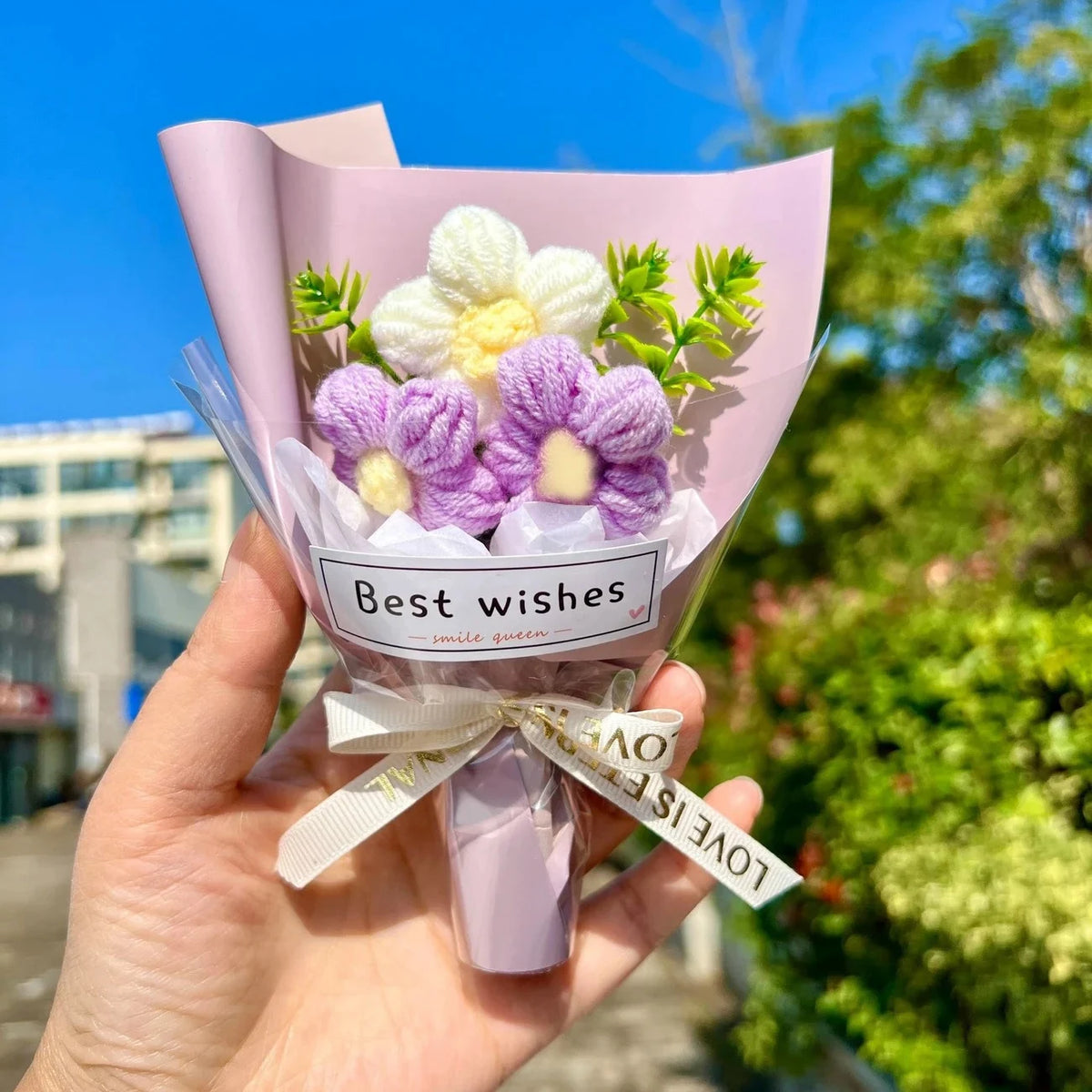 A hand holds a bouquet of handmade crochet flowers in a pink paper wrapper with a white ribbon and a love is in the bow tag.