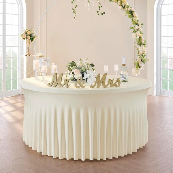 A round table draped with a floor-length, pleated ivory spandex tablecloth decorated with floral garlands and a wedding sign.