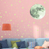 A pink wall decorated with light green stars and a large moon decal, next to a lampshade and blue furniture.