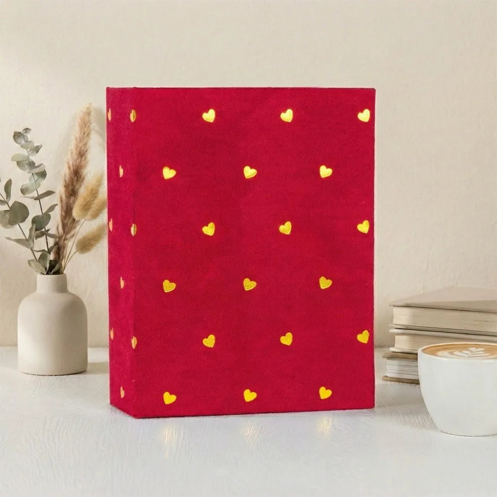 A red laminated photo album with gold heart patterns rests on a white surface next to a white mug.