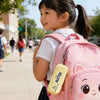 A young girl is carrying a pink plush butter dumpling squishy keychain backpack on a sunny day.
