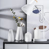 A white hand-frosted ceramic tabletop vase is displayed next to a modern desk lamp and some dried flowers.