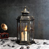 A vintage Nordic iron candle lantern with glass panes sits on a gray cloth surrounded by smooth dark stones.