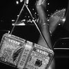 A hand holds a sparkling silver clutch bag and a pair of clear high heels against a dark background.