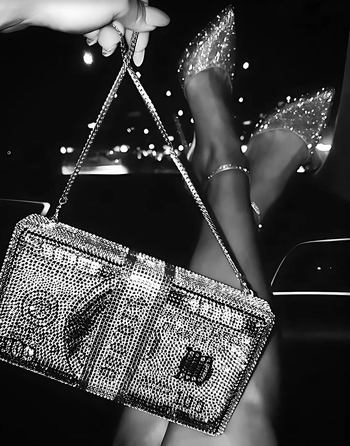 A hand holds a sparkling silver clutch bag and a pair of clear high heels against a dark background.
