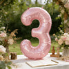 A large pink foil number balloon shaped like the number three is displayed outdoors among greenery and flowers.