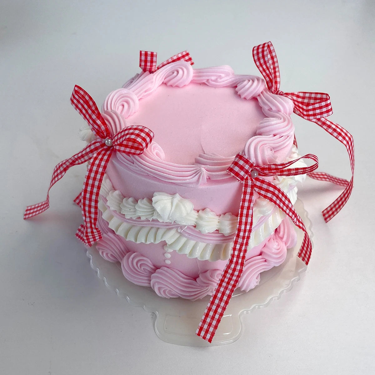 A pink decorative fake cake with piped frosting and red gingham ribbons on a clear stand.