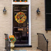 A black wooden door decorated with a large, colorful artificial flower wreath on a brick facade.