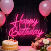 A pink neon sign spelling out Happy Birthday above a cake and floral arrangement.