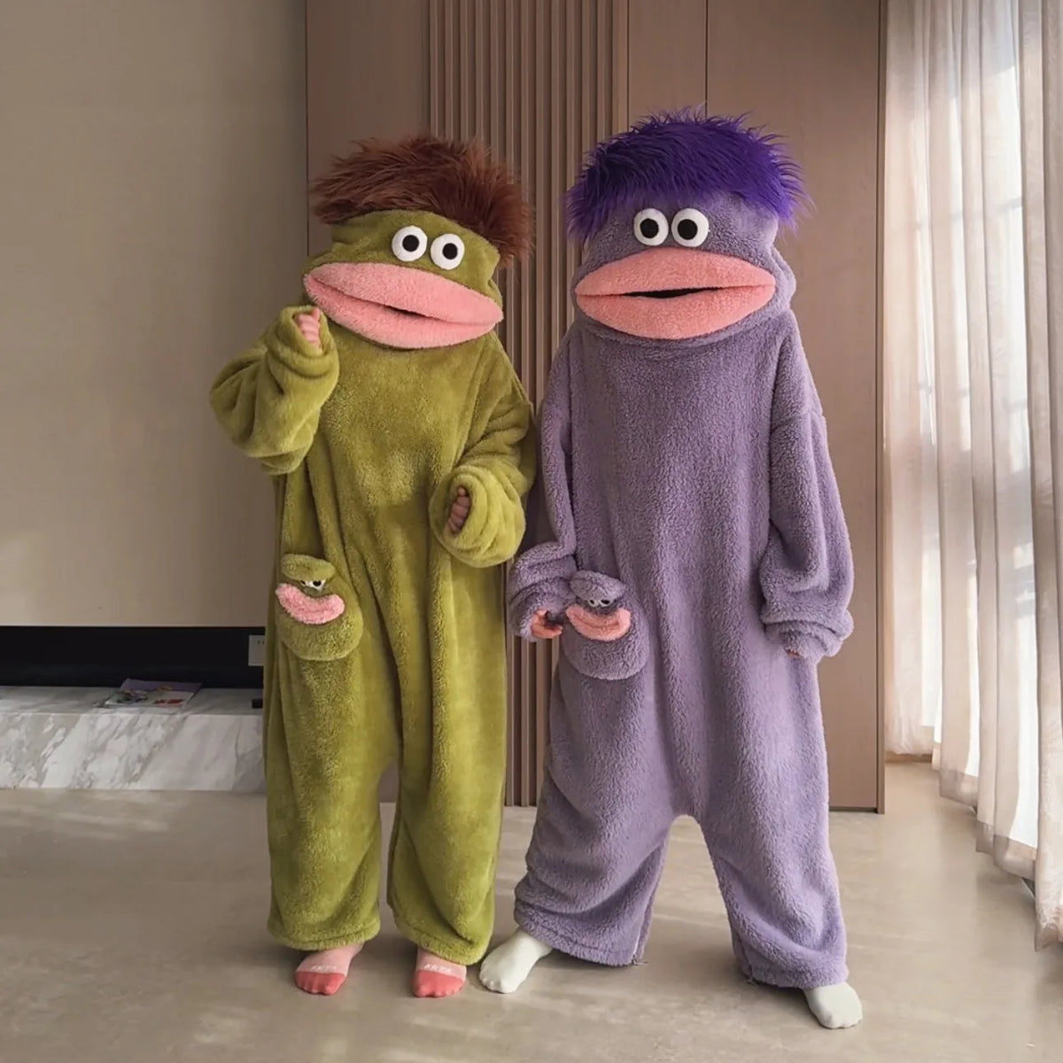 A pair of fuzzy green and purple monster onesie pajamas for couples sitting on a floor.