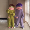 A pair of fuzzy green and purple monster onesie pajamas for couples sitting on a floor.